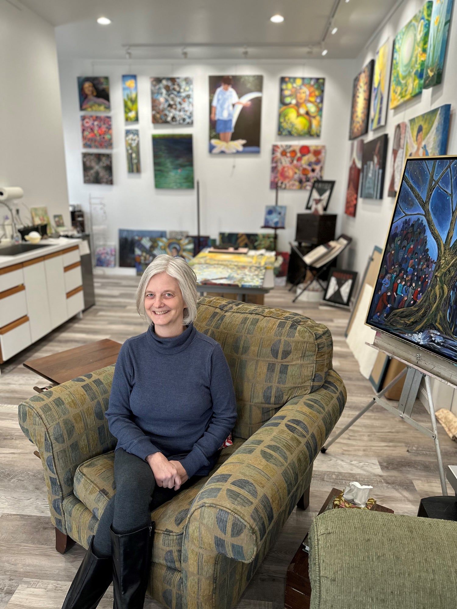 A woman sitting in an armchair in an artist studio. Paintings cover the walls and one on an easel behind her.