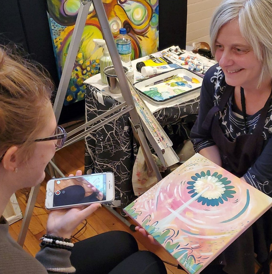 A profile of a woman on the right holding a cell phone. A woman on the right smiling and holding a painting while sitting in front of an easel with a table next to her full of paint.