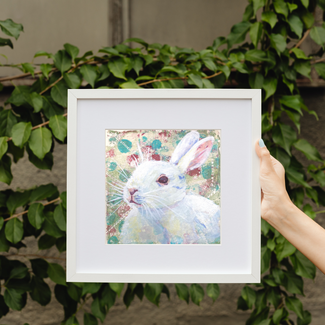 Original acrylic artwork by artist Tonya Henderson of a white rabbit in a frame held by a hand with green foliage in the background.