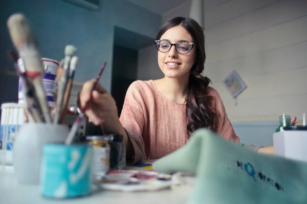 A woman in glasses with a paint brush in her hand. She is dipping it into water and has paint and brushes all around her.