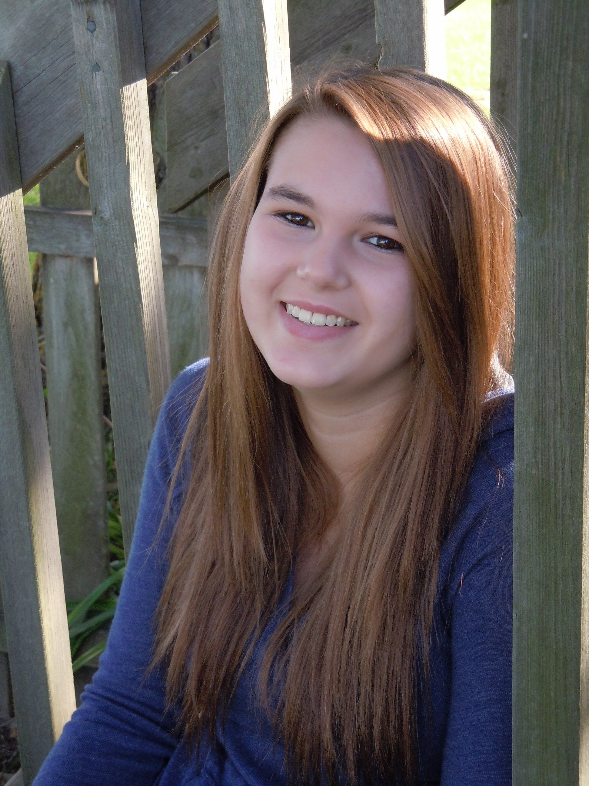 A photo of a young lady dressed in royal blue. She is on a deck of aged wood, has long brown hair, and is smiling.