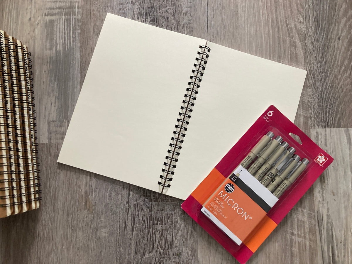 An open spiral-bound notebook with blank cream pages lies on a gray wood-textured surface, with a package of Micron gel pens placed on top and a stack of similar notebooks on the right side.
