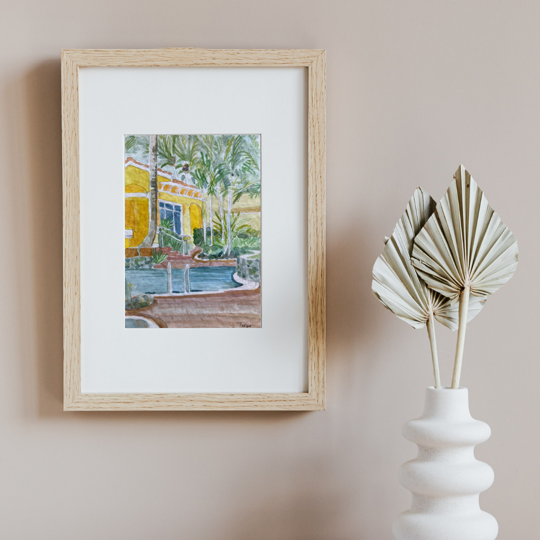 Framed original watercolor artwork of a yellow bathhouse with palm trees on a beige wall, next to a white vase with dried leaves. Artist Tonya Henderson.