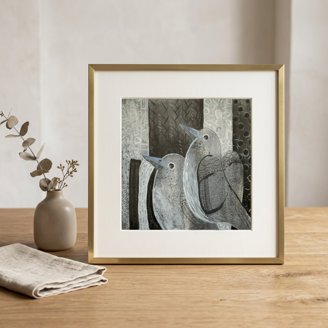 Framed artwork of birds on a wooden table with a vase and cloth in the background