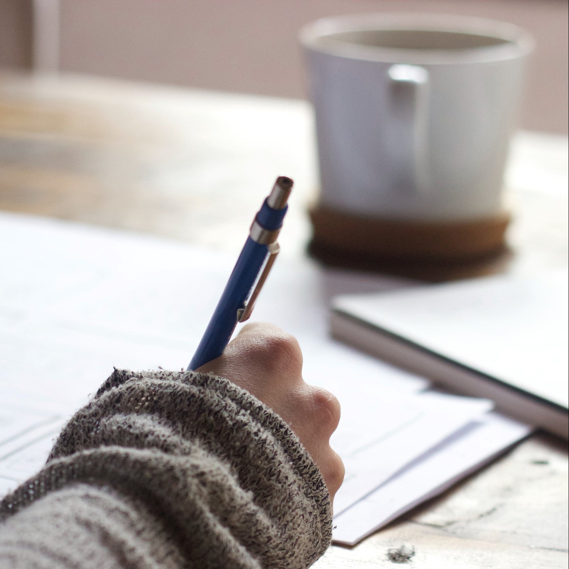 Hand holding a pen over a piece of paper on a desk with a mug in the background