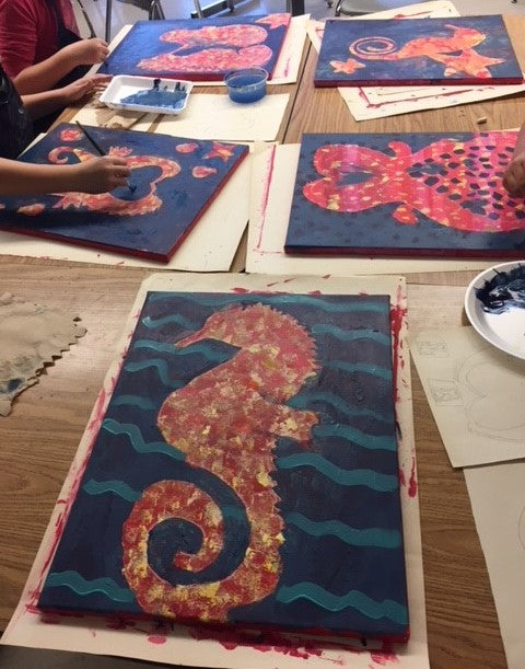 Students painting artistic seahorse designs on canvases with paint supplies on a table.