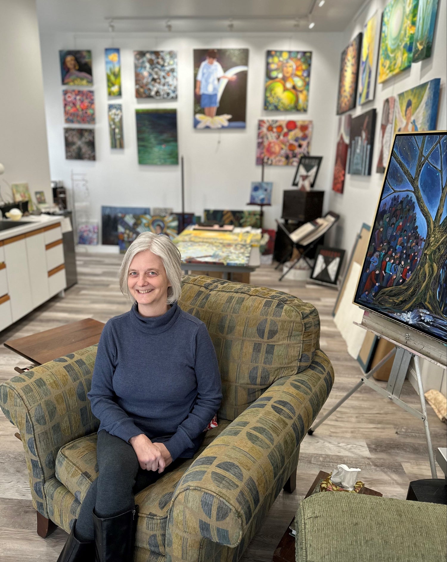 Artist Tonya Henderson sitting in an armchair in her artist studio. Paintings cover the walls and one on an easel behind her.