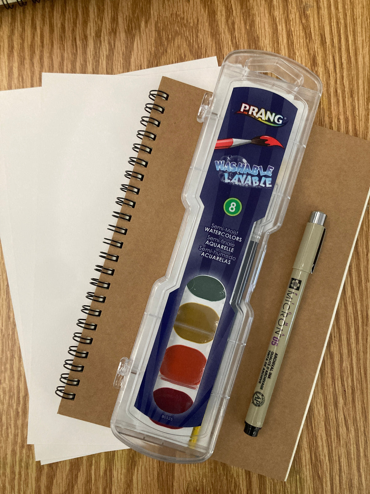 A Frang watercolor paint set in a clear plastic case, shown with a beige pen and sketchbooks on a wooden desk.
