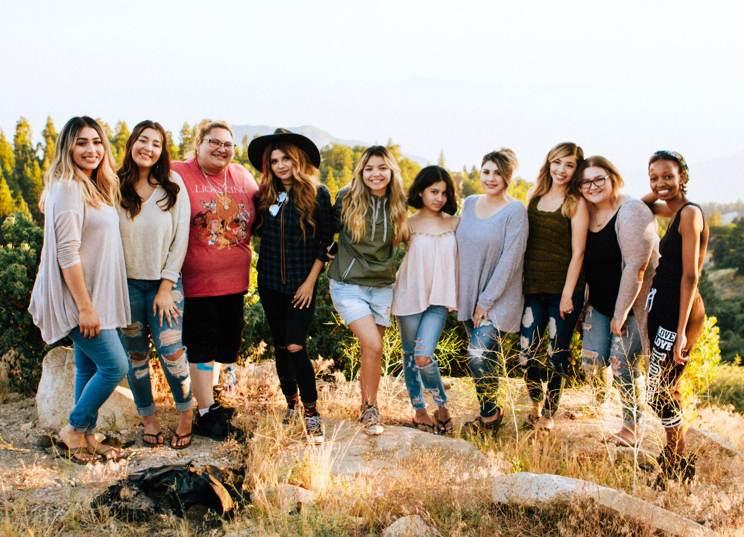 A group of woman standing in a row at the top of a rocky mountain in summertime clothing. 